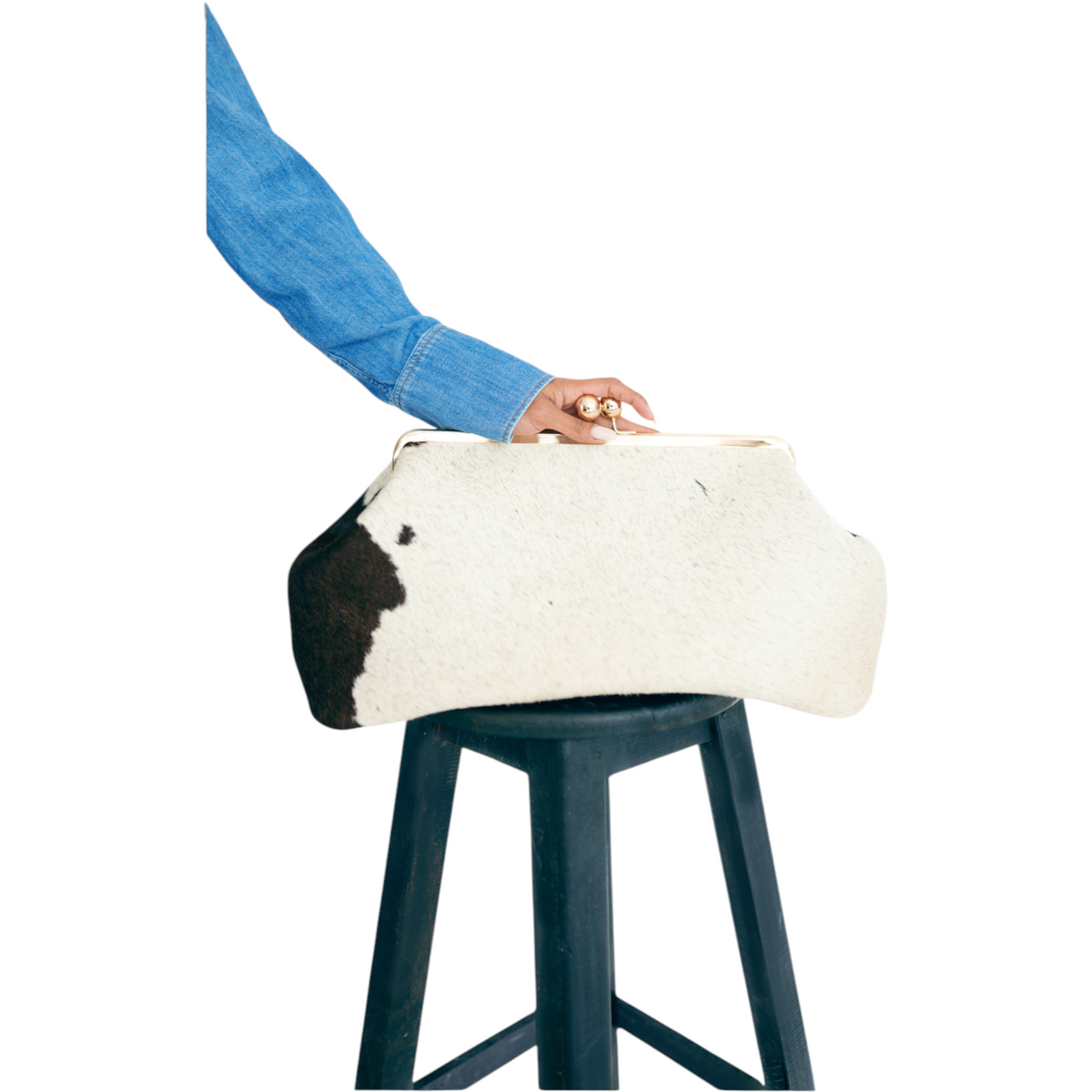 Jesalin Sette cow print leather pouch with gold clasp held by a person wearing a denim shirt on a black stool