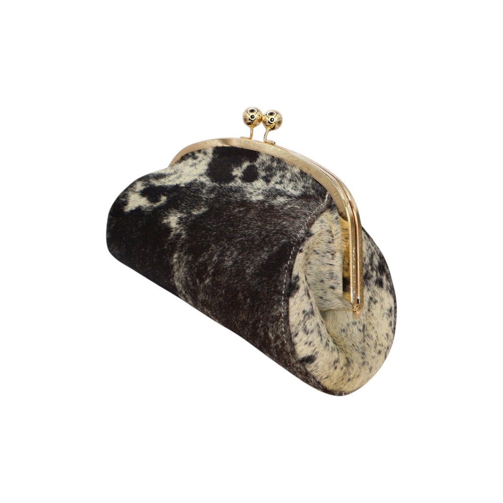 Clutch rígido Jesalin Aluna en piel bovina con estampado de vaca, marco metálico dorado y cierre tipo kiss lock.
